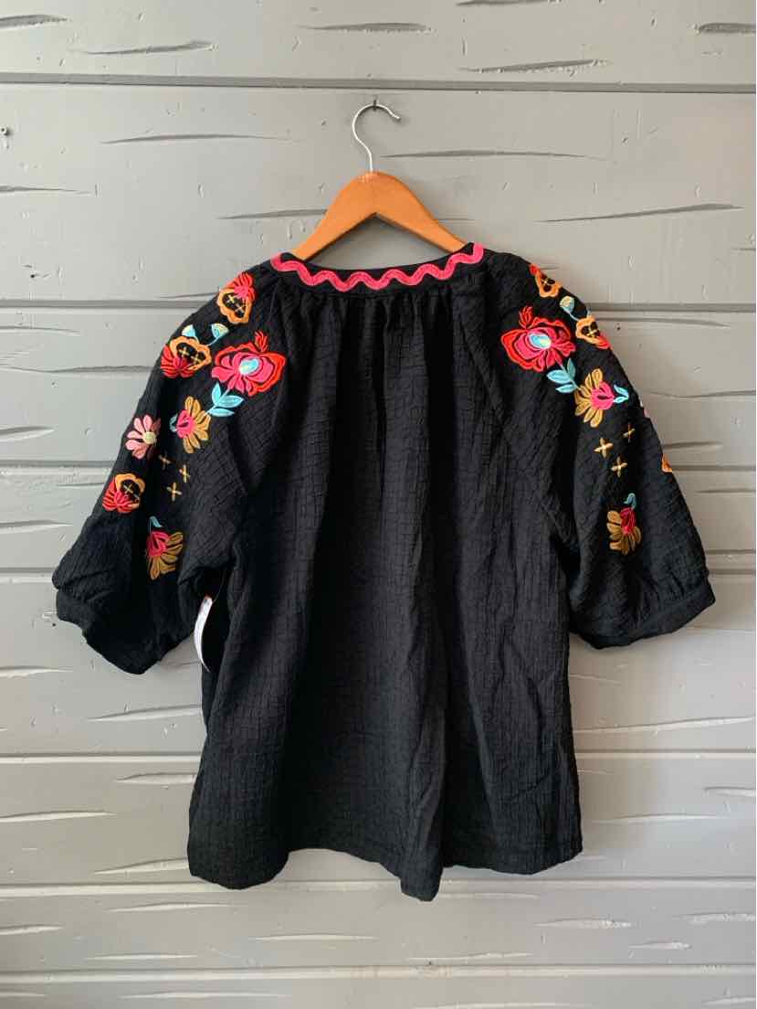 Black loose-fit blouse with wide sleeves, colorful floral embroidery on shoulders, and pink scalloped neckline trim.