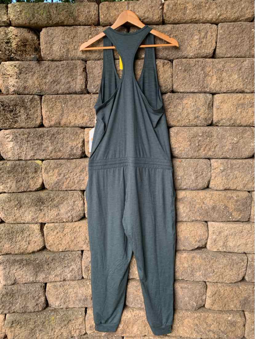 Gray sleeveless jumpsuit with racerback design and tapered legs hanging on wooden hanger against stone wall.