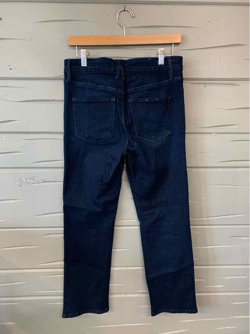 Dark blue denim jeans with straight legs and rear patch pockets hanging on wooden hanger against white wooden wall.