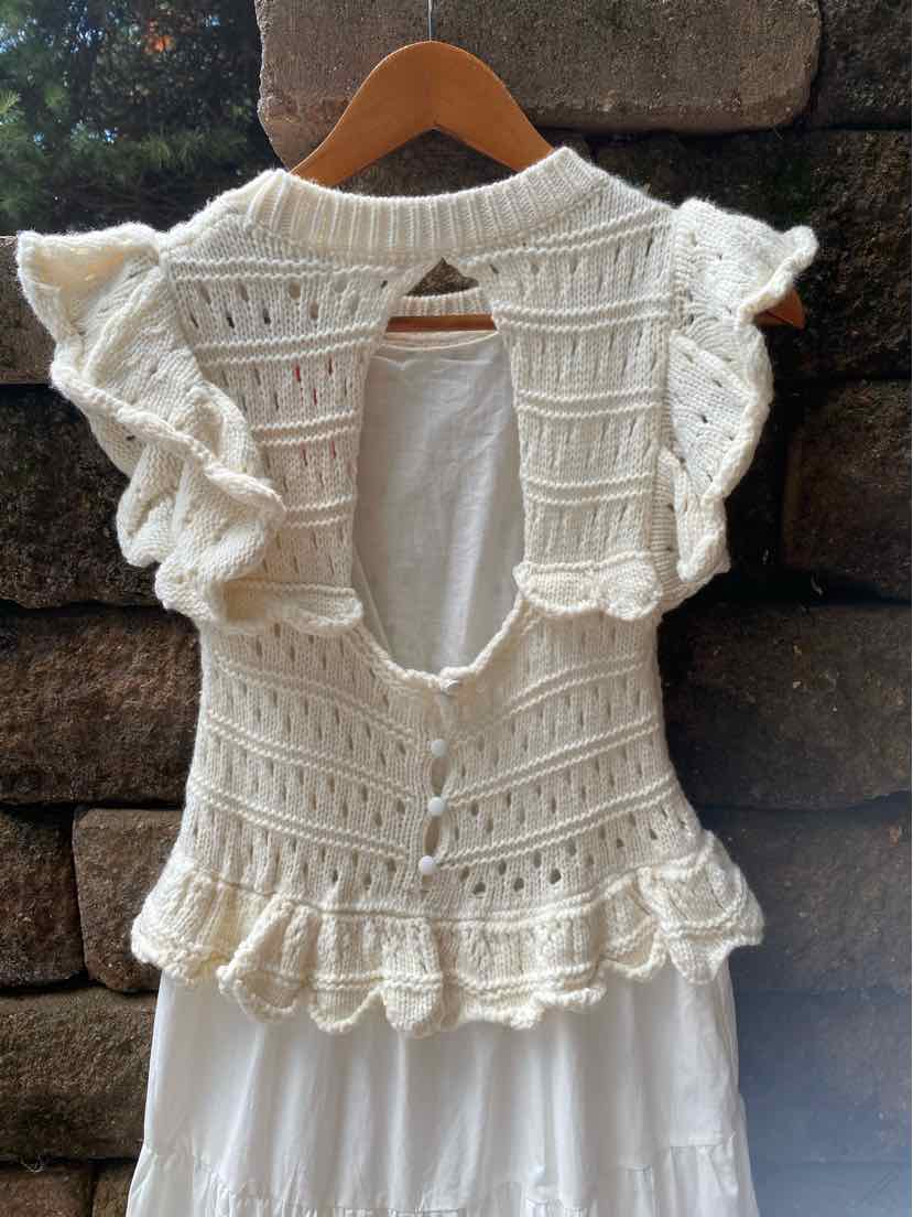 White knitted sleeveless vest with ruffled shoulders and peplum hem, open back with button closure.