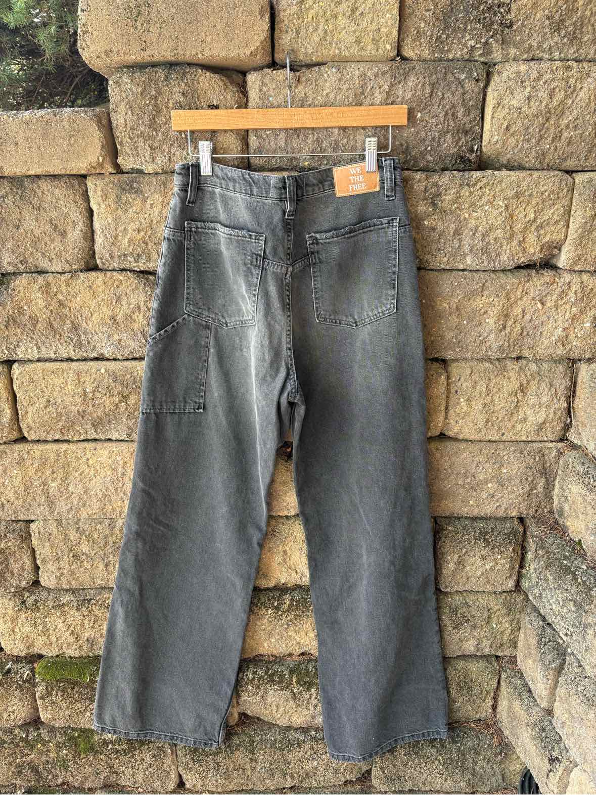 Charcoal gray denim jeans with a faded wash, straight legs, and a tan waistband patch hanging against a stone wall.