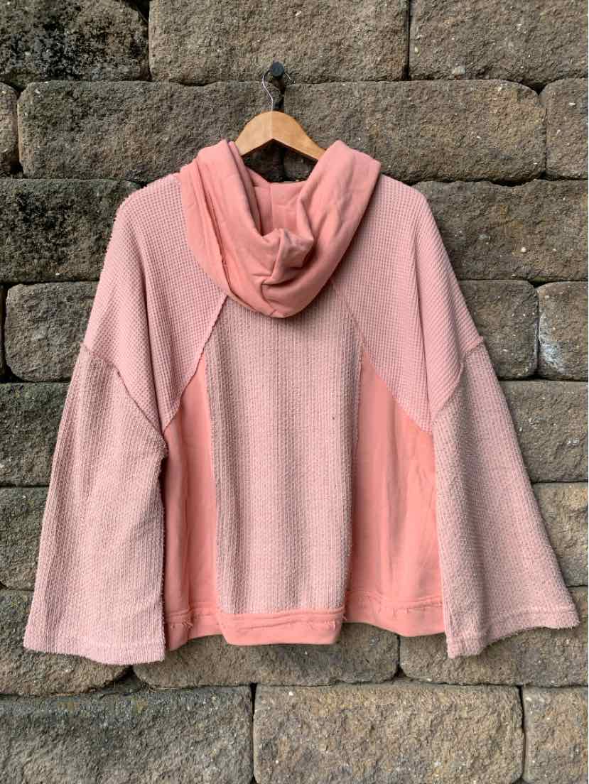 Dusty rose pullover hoodie with a soft fleece texture, hood, and wide sleeves hanging against a stone wall.