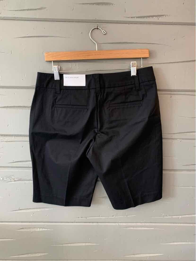 Black tailored knee-length shorts with back flap pockets hanging on wooden hanger against light gray wooden wall.
