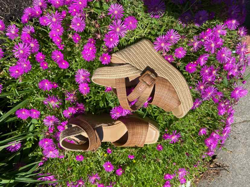 Tan woven wedge sandals with ankle straps and open toes on green grass surrounded by purple flowers.