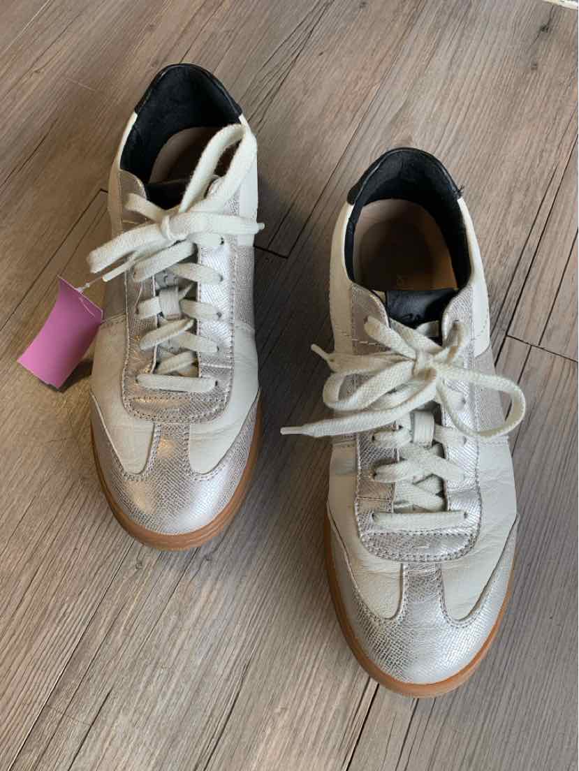 White leather sneakers with beige detailing, white laces, and gum soles on a wooden floor.