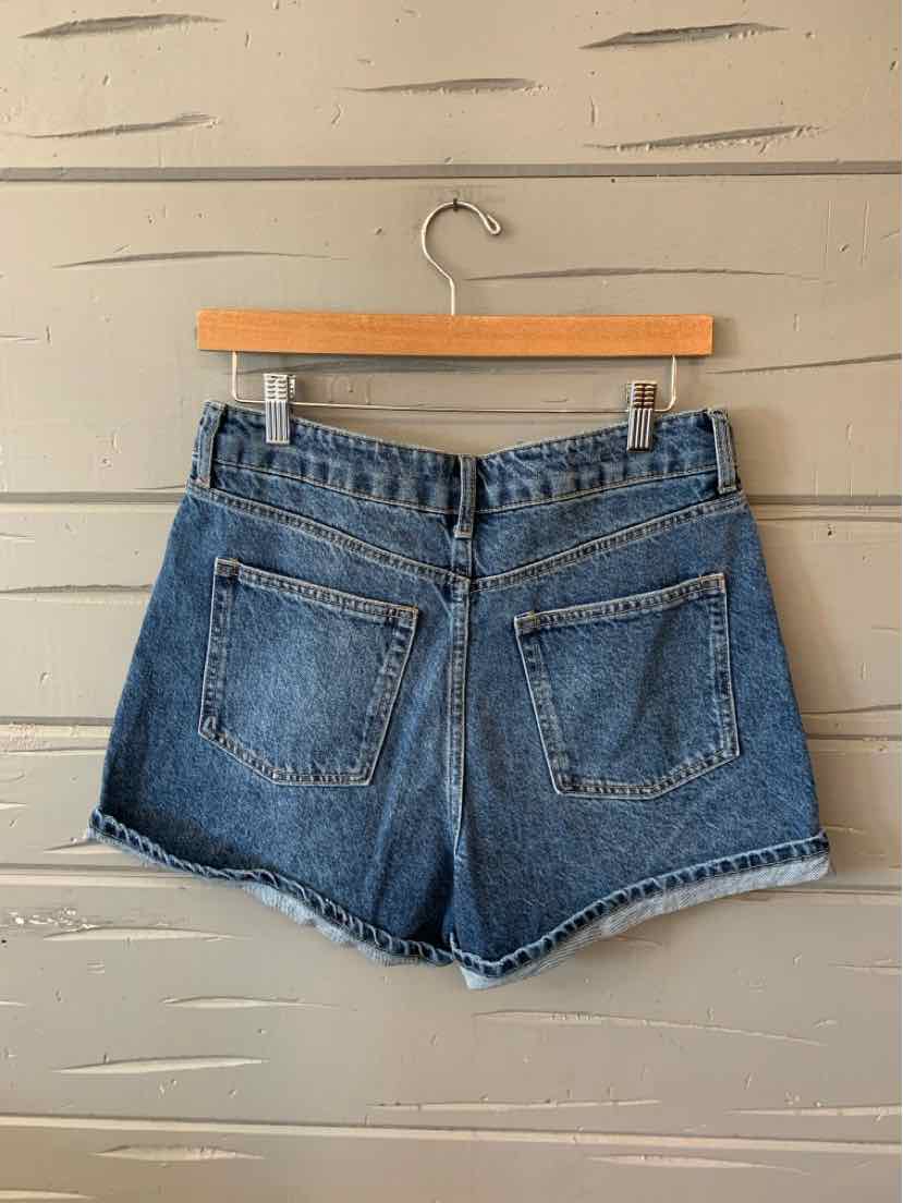 Blue denim shorts with a high waist and two back pockets hanging on a wooden hanger against a beige wooden wall.