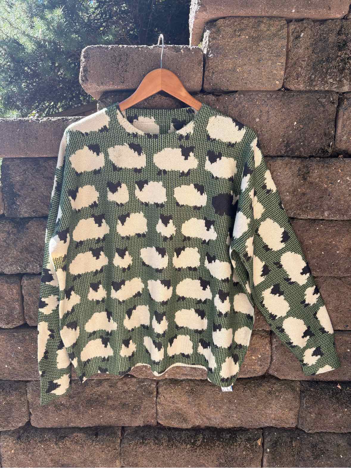 Olive green knit sweater with repeating cream sheep pattern and ribbed cuffs, hanging from a wooden hanger.