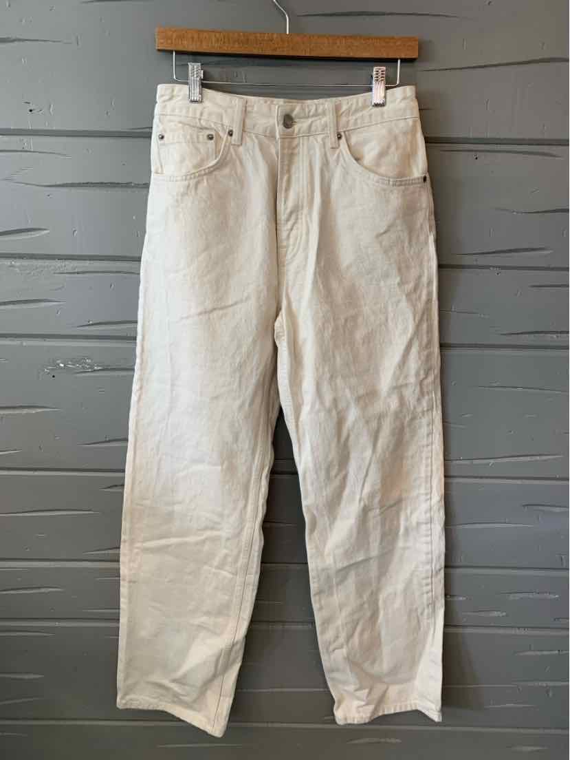 Light beige straight-leg jeans with five pockets and belt loops hanging on wooden hanger against gray wooden wall.