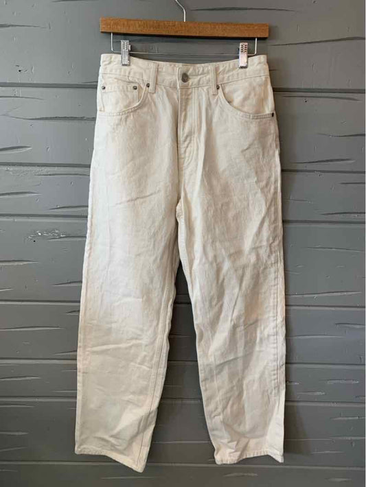Light beige straight-leg jeans with five pockets and belt loops hanging on wooden hanger against gray wooden wall.