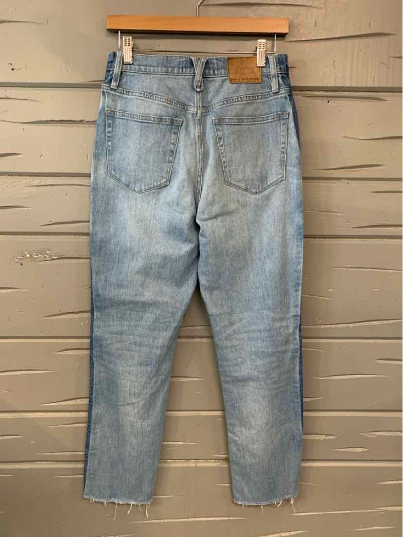 Light blue denim jeans with frayed hems and two back pockets hanging on wooden hanger against a beige wooden wall.