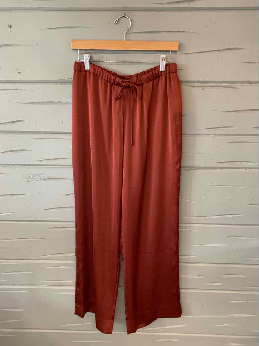 Brick red wide-leg pants with drawstring waist hanging on wooden hanger against rustic white wooden wall.