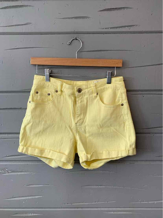 Yellow denim shorts with front pockets and button closure on wooden hanger against gray wooden background.