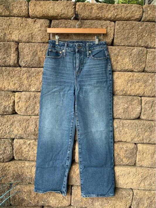 High-waisted blue denim jeans with a straight leg and front pockets hanging on a wooden hanger against a stone wall.