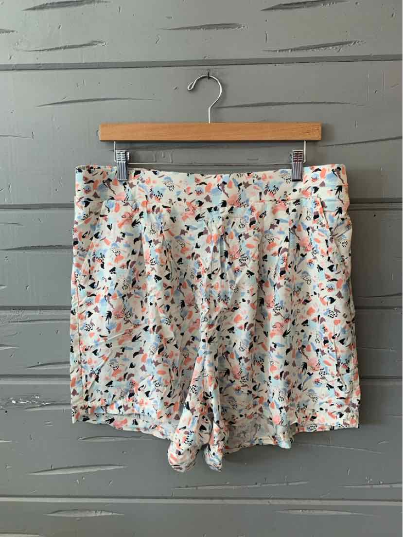 White shorts with an all-over abstract floral pattern in blue, orange, and black, hanging on wooden hanger.