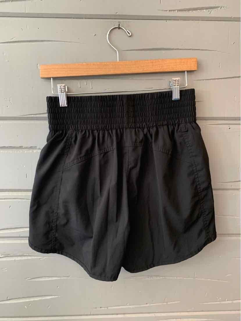 Black athletic shorts with wide elastic smocked waistband and loose fit, hanging on wooden hanger against gray wooden wall.
