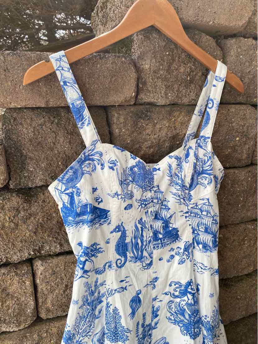 White dress with blue floral toile print, fitted bodice, and wide shoulder straps on wooden hanger.