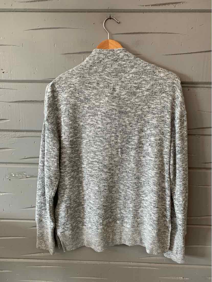 Light gray knit sweater with long sleeves and ribbed cuffs hanging on wooden hanger against wood panel background.