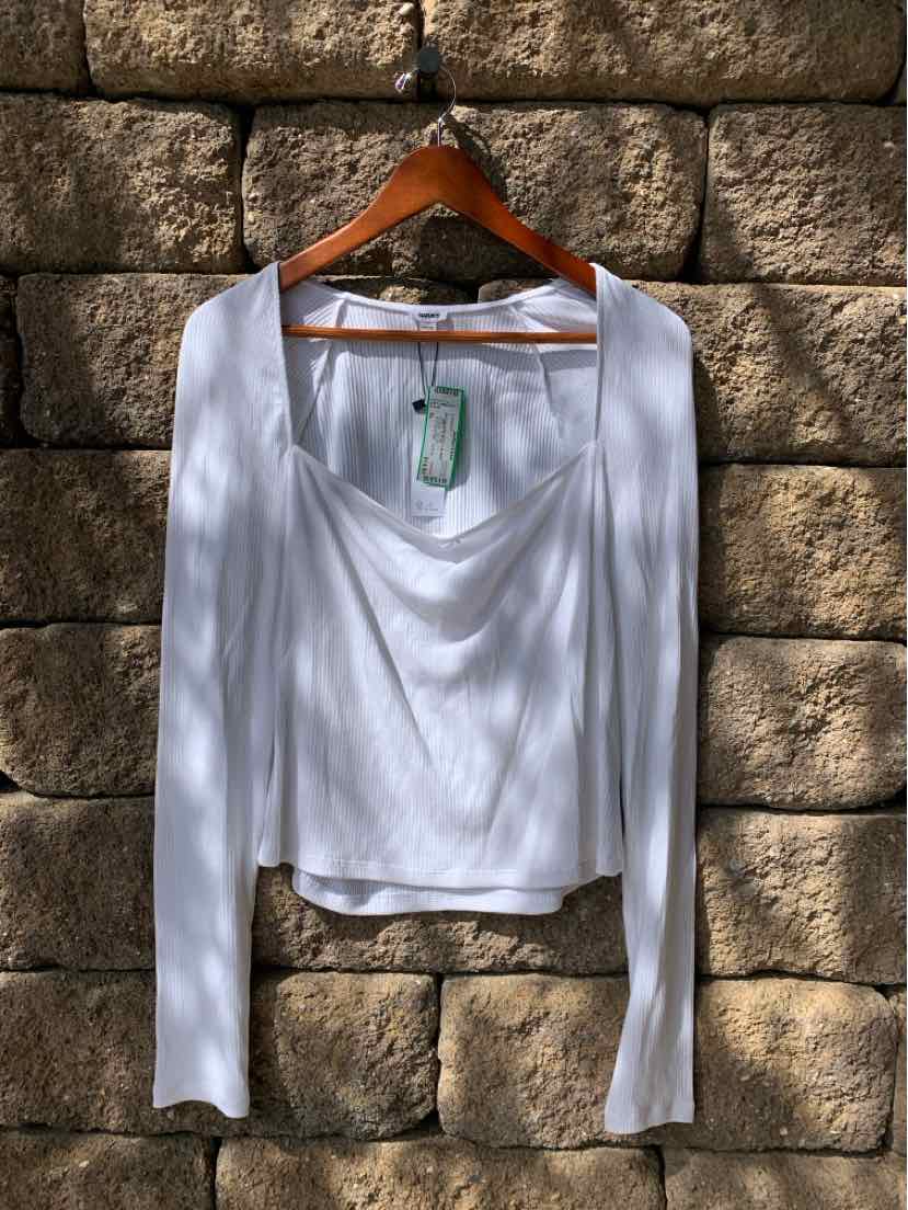 White long-sleeve top with draped neckline and fitted silhouette hanging on wooden hanger against stone wall.