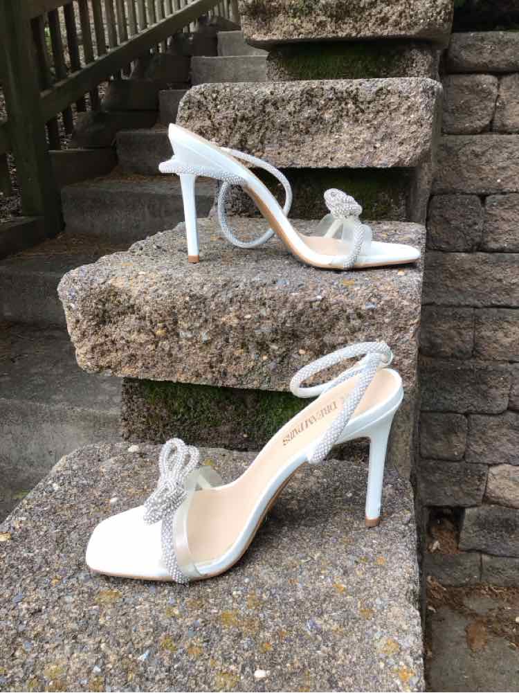 White high-heeled open-toe sandals with ankle straps and rhinestone bow accents on stone steps.