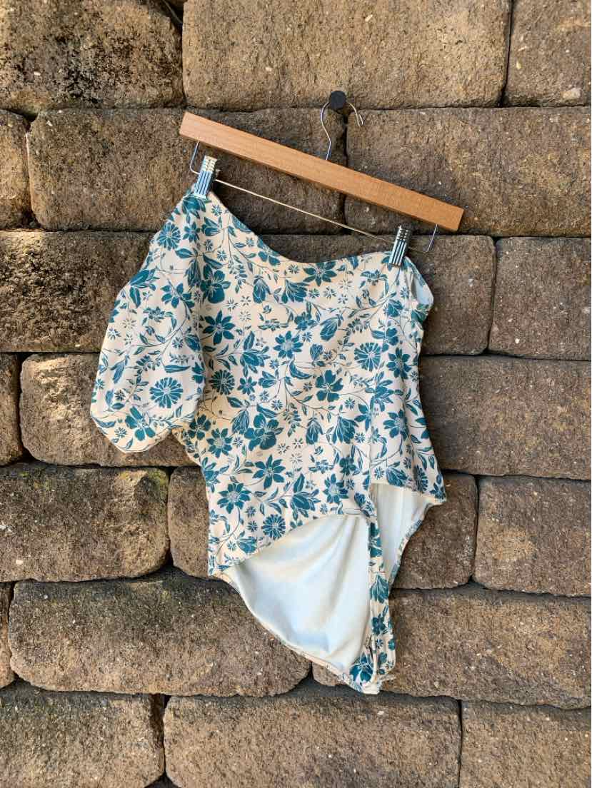 White one-shoulder swimsuit with blue floral print and elasticized waist detail hanging on wooden hanger against stone wall.