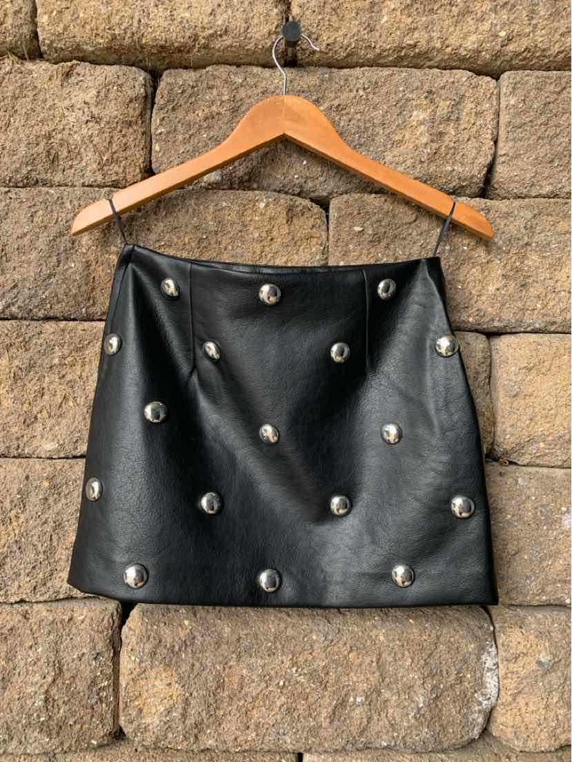 Black faux leather mini skirt with silver stud embellishments hanging on wooden hanger against stone wall.