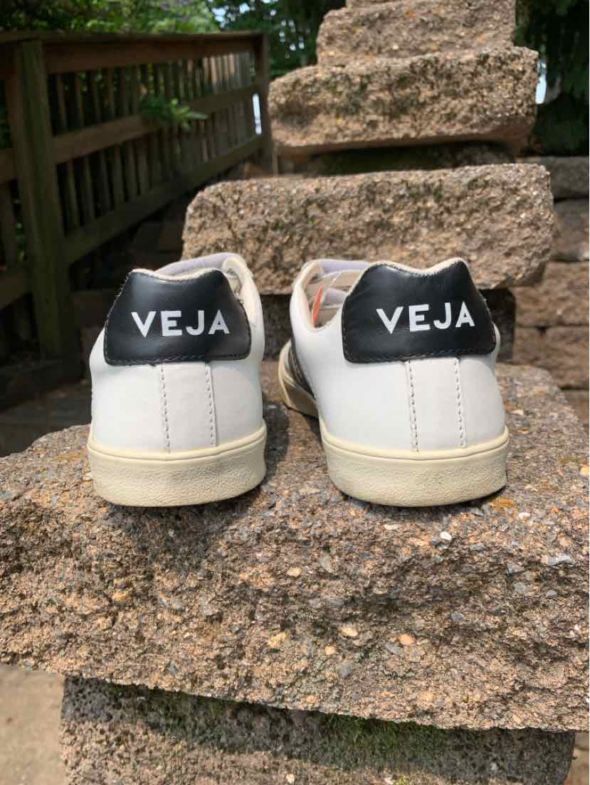 Pair of white low-top sneakers with black heel tabs featuring "VEJA" branding in white letters, off-white soles, and a.
