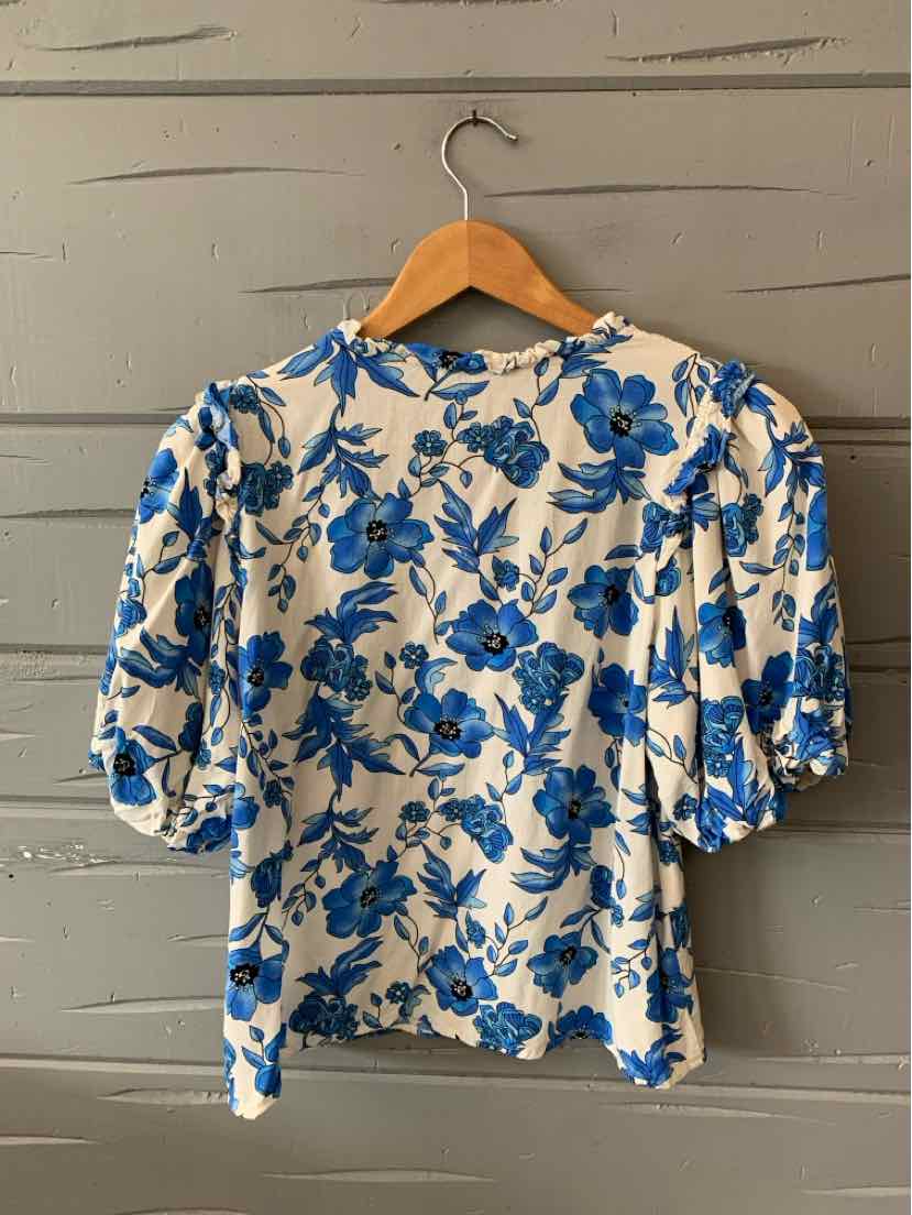 White short-sleeve blouse with blue floral print and slight puff sleeves hanging on wooden hanger.