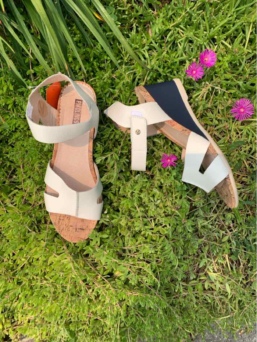 Beige leather sandals with adjustable ankle straps and open toes on green grass and purple flowers.