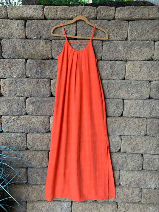 Sleeveless bright orange maxi dress with thin straps and flowing fabric on wooden hanger against stone wall.