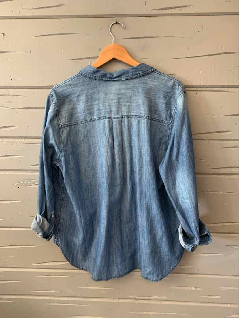 Light blue denim button-up shirt with long sleeves rolled up, displayed hanging from a wooden hanger against a textured wall.